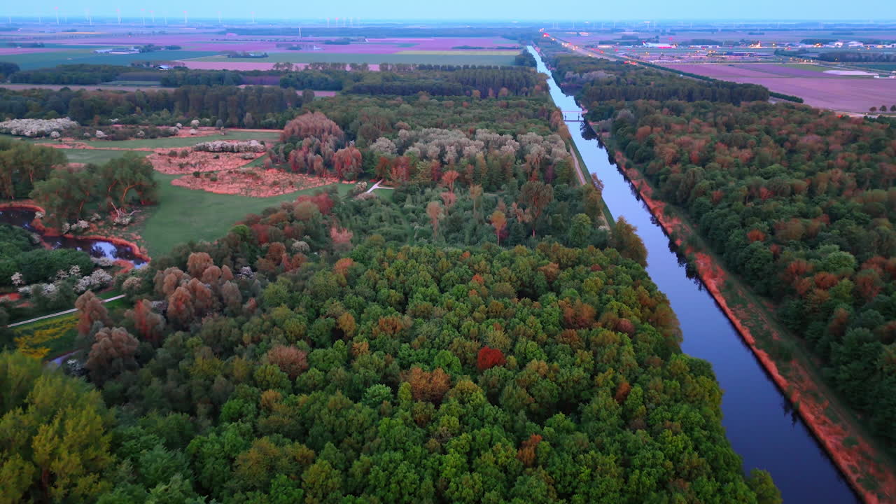 Amazing green lush forest crossed by the canal. Agricultural fields at backdrop. Drone flight above the countryside.