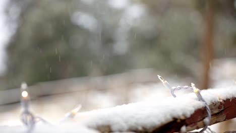 Close-up-of-white-Christmas-lights-on-a-snowy-deck-railing-with-pine-trees-in-the-background-and-snow-falling