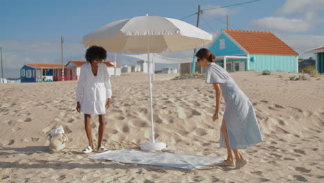 Girlfriends-spread-picnic-blanket-on-sandy-beach.-Two-girls-enjoying-summer-day