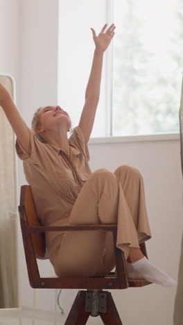 happy blonde woman with ponytail spins on wooden chair putting hands up with joy against mirror in contemporary fitting room at home slow motion