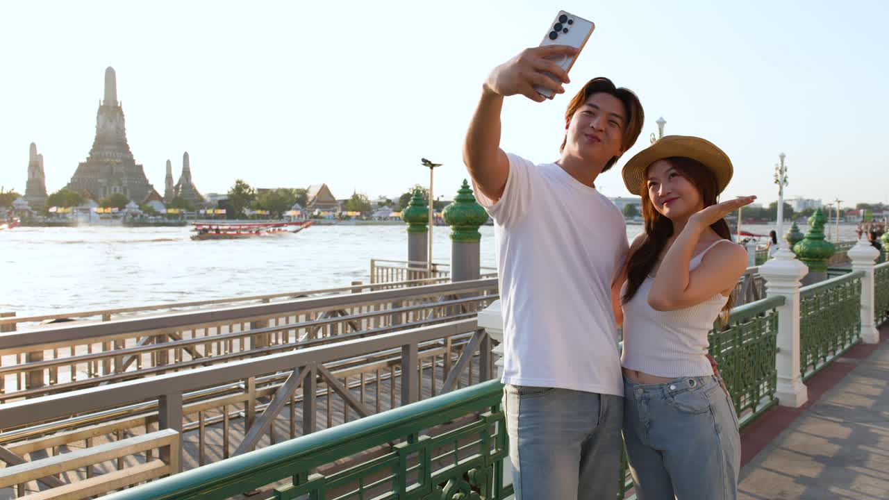 Coppia sorridente si scatta un selfie sulla passeggiata lungo il fiume, luce dorata del tramonto, sfondo con un tempio iconico