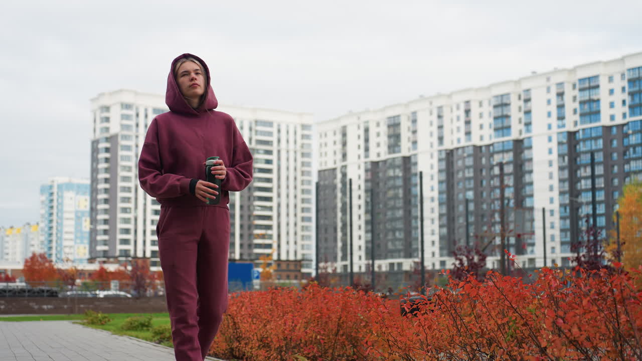 Jogger walking in burgundy hoodie and matching sweatpants clutching green water bottle while strolling urban pavement lined with red shrubs and spring flowers beneath tall modern apartment buildings