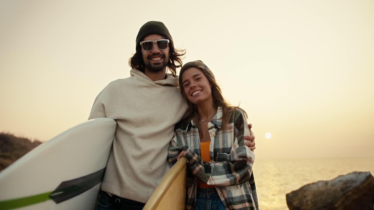 retrato de una pareja feliz, un hombre moreno con gafas de sol y una sudadera blanca se encuentra con su novia rubia en una camisa a cuadros y sostienen tablas de surf cerca del mar contra el telón de fondo de una flor brillante soleada por la mañana en otoño