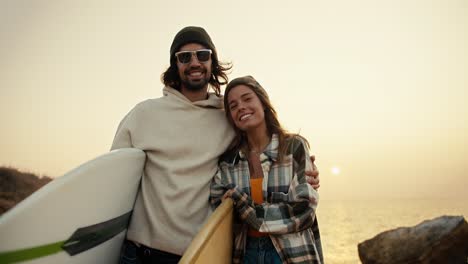 portrait of a happy couple, a brunette man in sunglasses and a white sweatshirt stands with his blonde girlfriend in a plaid shirt and they hold surfboards near the sea against the backdrop of a sunny bright bloom in the morning in autumn