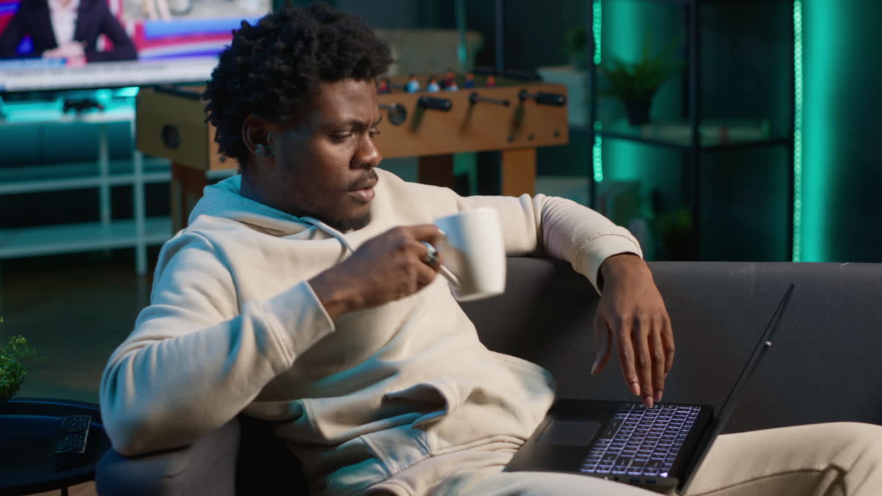 Positive african american man multitasking on a laptop in stylish loft, fully connected to the internet while managing his freelance work and remote responsibilities. Browsing web at home. Camera A.