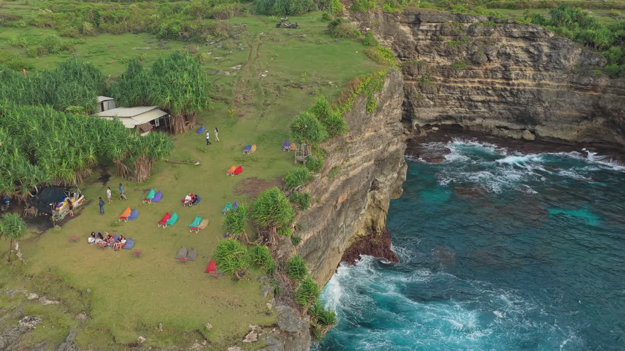 Incredibile vista aerea di persone che si rilassano su pouf colorati in cima a una scogliera verde che si affaccia sulle onde turchesi dell'oceano a Bali.