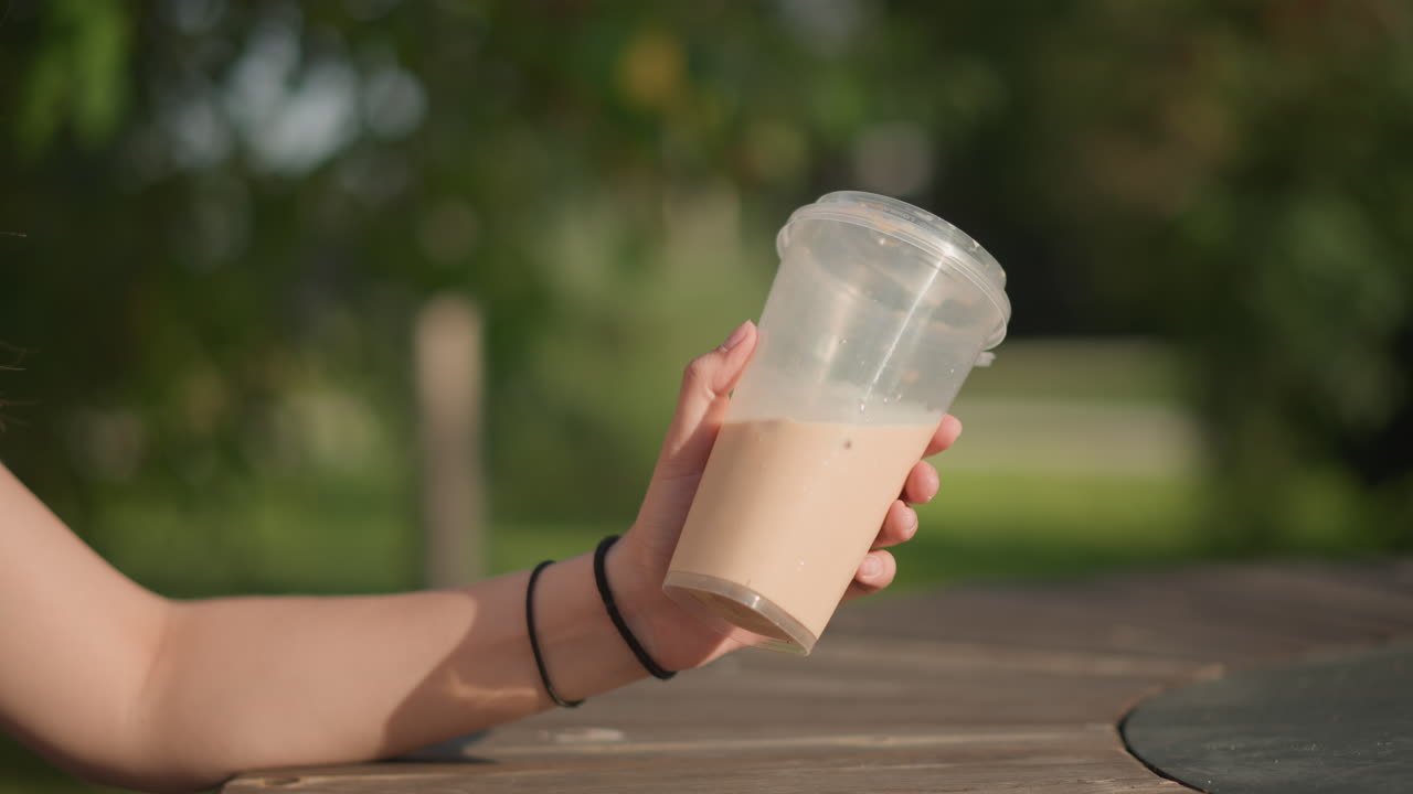 Giovane donna che tiene in mano un latte macchiato freddo seduta su una panchina del parco sotto una luce dorata soffusa, creatrice di contenuti che inquadra uno scatto di lifestyle, modella che posa per un primo piano, studentessa che si ferma per un sorso, venditore che assaggia, amico che fa un brindisi