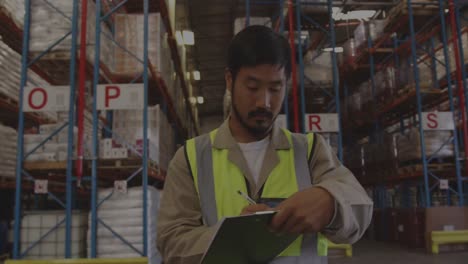 Animation-of-graphs,-loading-circles-and-bars,-smiling-asian-man-writing-in-notepad-at-warehouse