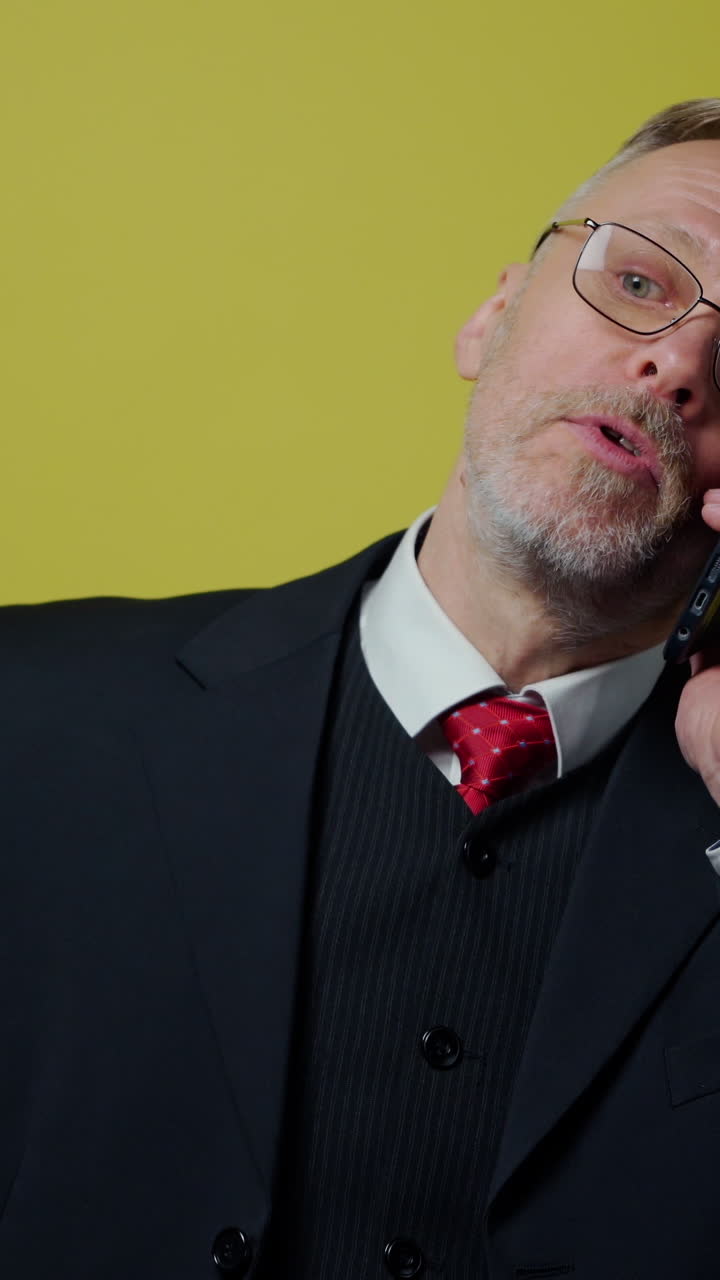 Nervous middle aged businessman with a cellphone. Mature man in dark suit talking on the phone and giving instructions to somebody on yellow background. Vertical video