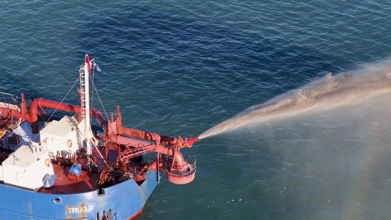 A dredging vessel pumps sand into the ocean, creating a rainbow effect under bright sunlight