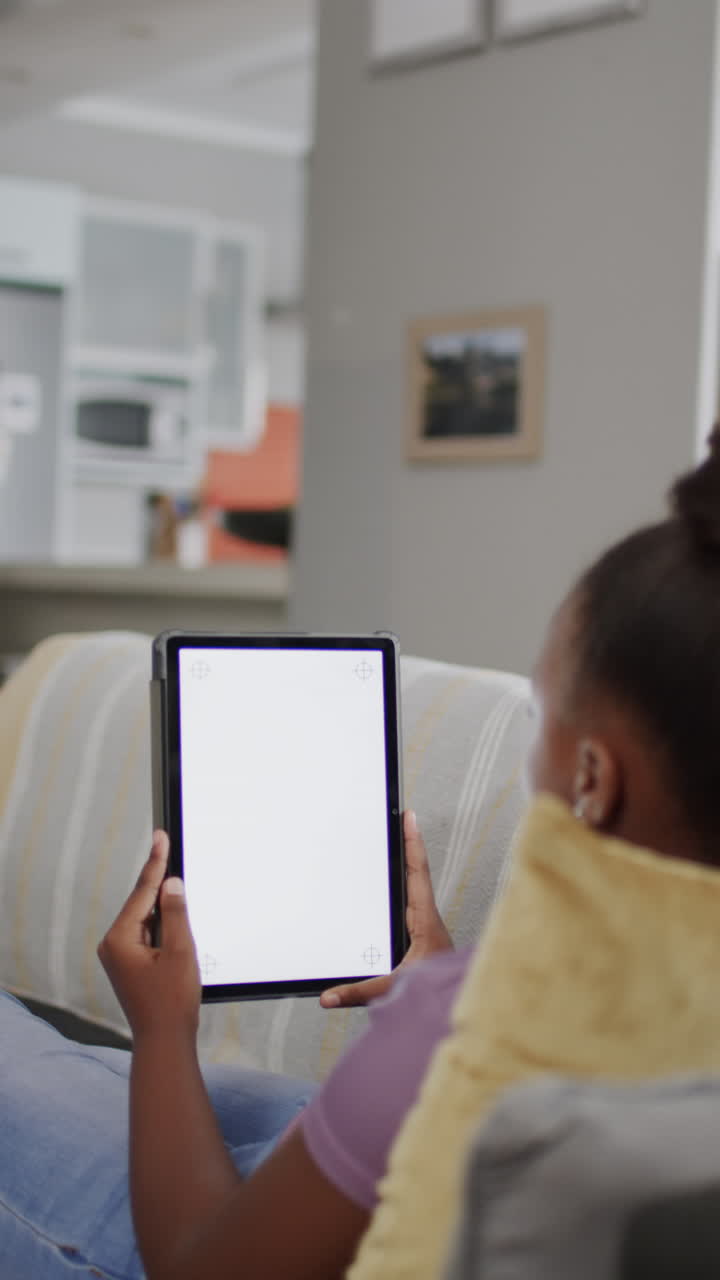 Vertical video of african american girl sitting on sofa, using tablet with copy space, slow motion