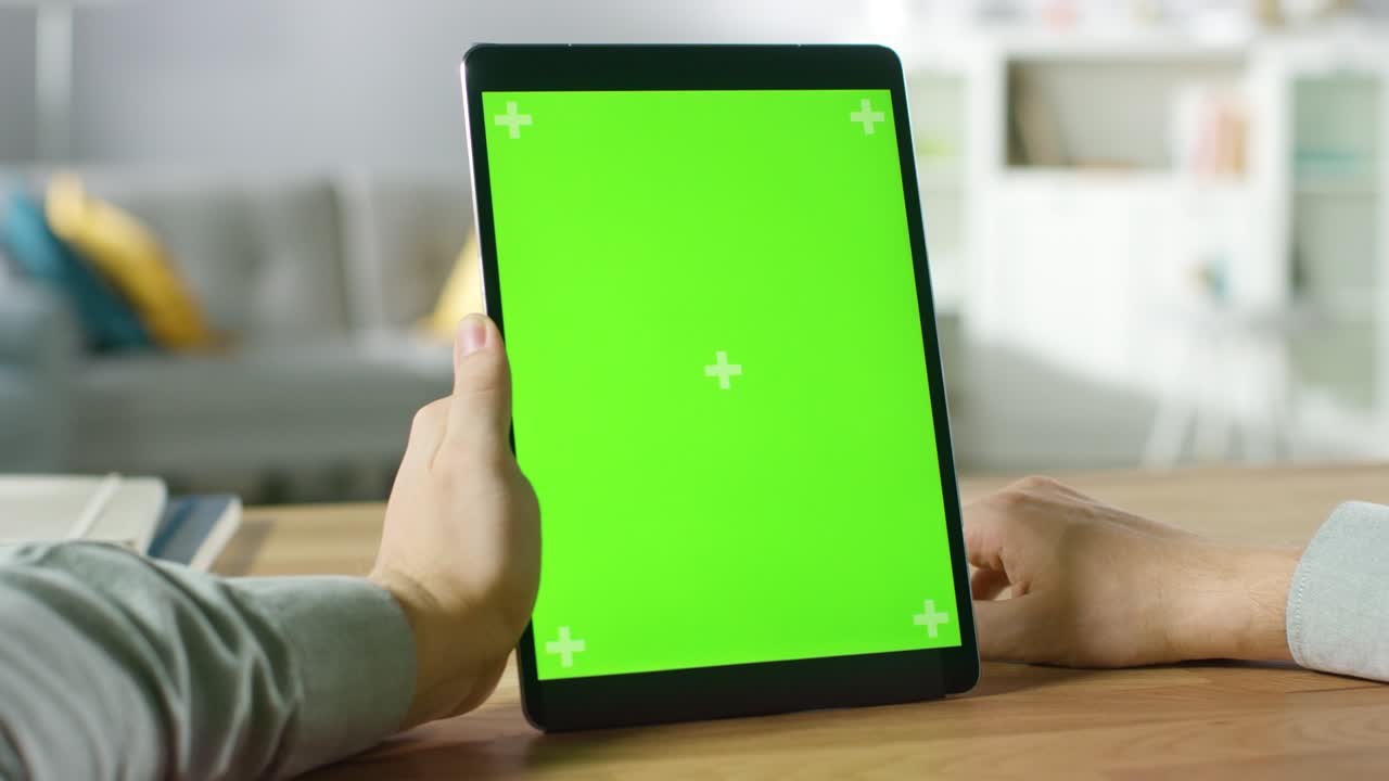 Nærbillede af mand, der bruger håndbevægelser på grøn mock-up skærm tablet i portrættilstand, mens han sidder ved sit skrivebord. I baggrunden hyggelig stue