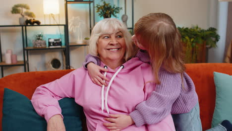 Cheerful-loving-small-girl-granddaughter-embracing-kissing-happy-grandmother-sitting-on-home-sofa