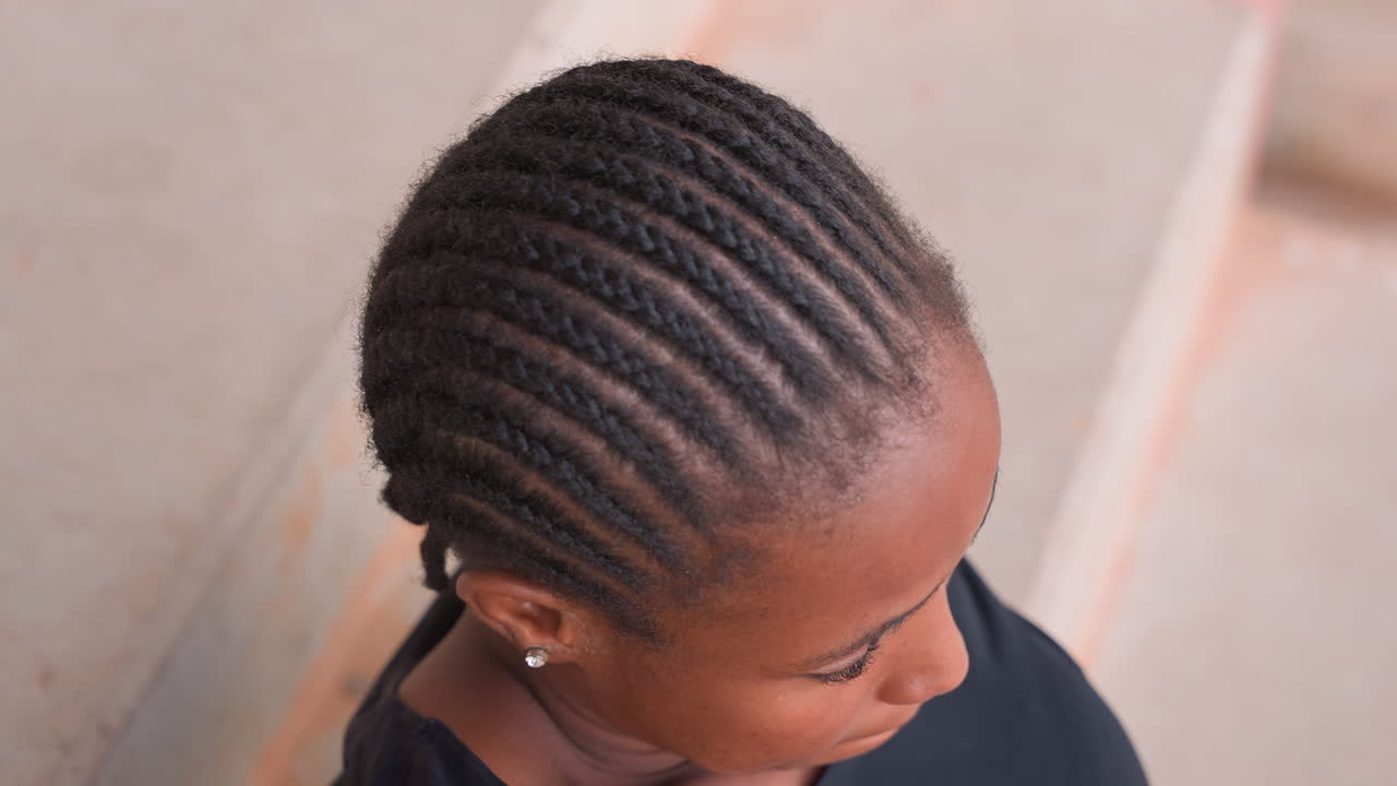 Portrait de femme noire, yeux fermés, respirant sereinement sur des marches en béton, capturant les cheveux tressés, la courbure du cou et une lente inspiration. Moments authentiques de relaxation et de lâcher-prise, peau baignée de soleil et humeur calme.