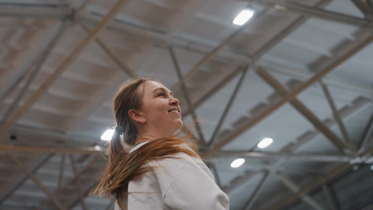 Young woman on indoor badminton court wearing white sweatshirt and ponytail, looking up with focused smile while preparing overhead serve during training session under steel roof trusses and bright