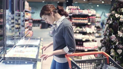 Young female customer comes up to a freezer, compares price tags and takes a pack of dumplings. Girl puts a pack in shopping