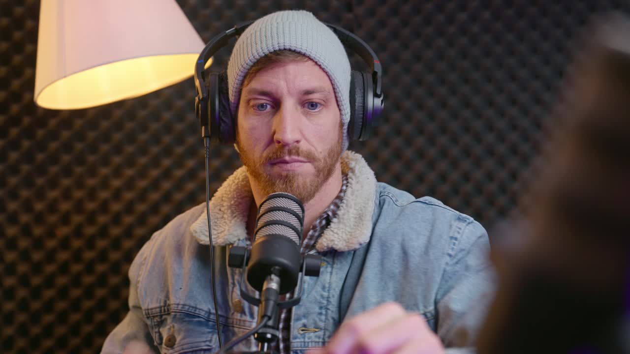 Cool hipster podcast host or radio announcer prepares for recording session in studio. Professional sound booth. Man with knit hat start live show streaming online. Setting up gear and talks excitedly
