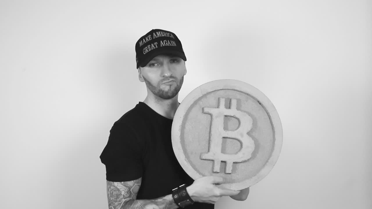 Grayscale footage of a tattooed man with a dark black MAGA or Make America Great Again Trump presidential hat lifting up a concrete Bitcoin cryptocurrency statue or sculpture and nods with approval.