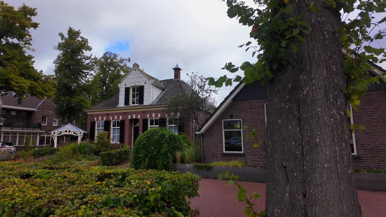 Traditional brick house with white gable, black shutters, and garden framed by a large tree in Ommen. Location: Ommen, Netherlands Ommen, Nederland