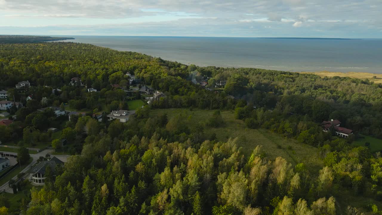 Aerial drone video of a forest covered cliff bank shoreline at Tabasalu by Baltic blue colored ocean sea water at summer sunny day. Private houses with smoking chimneys visible below, green gardens