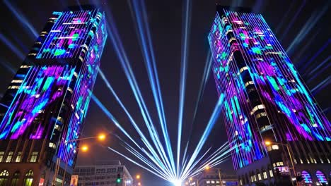 vibrant laser projections casting geometric patterns across metropolitan skyline, illuminating tall buildings with intense light streams converging centrally during nighttime cityscape