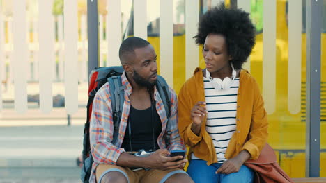 african american young happy man and woman travellers sitting at bus stop talking to plan a route