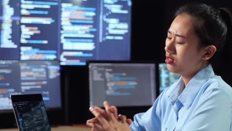 close up side view of asian female programmer stretching while writing code by a laptop using multiple monitors showing database on terminal window desktops in the office