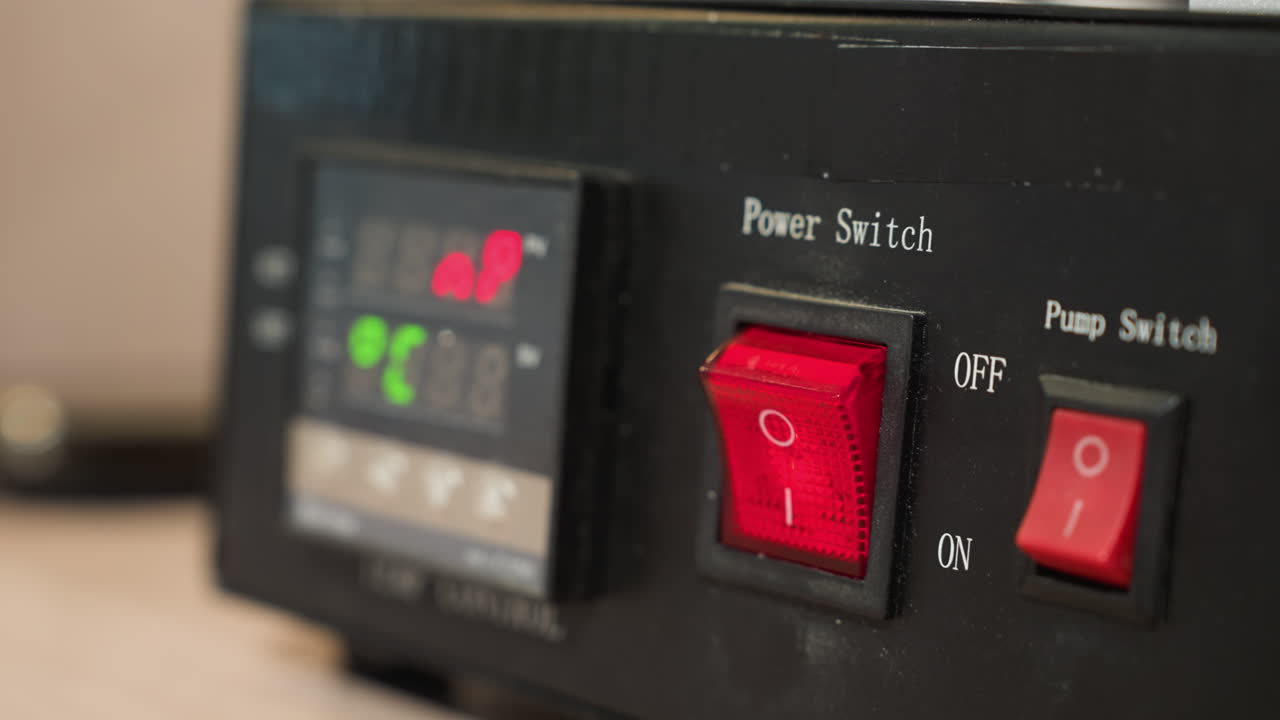 una vista de cerca de la mano con guantes de un técnico presionando el interruptor de energía rojo en un dispositivo de control de temperatura negra