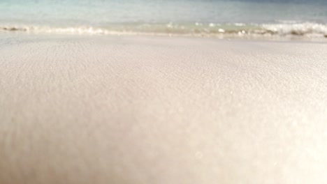 Turquoise-waves-gently-lap-against-the-wet-and-sparkling-golden-sand-in-summer,-coming-into-focus-as-the-camera-sits-at-ground-level-view-with-a-shallow-depth-of-field