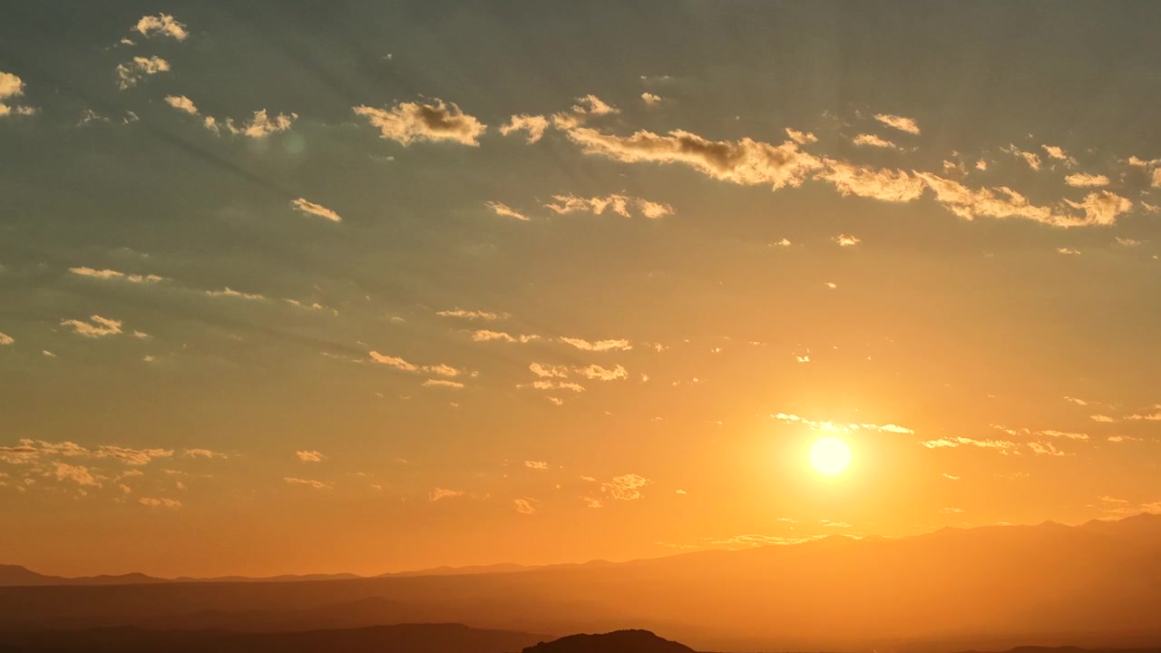 4K drone timelapse of a golden sunrise over a distant mountain range. Brilliant sunbeams shift dynamically with morning clouds, illuminating the vast and rugged landscape below.