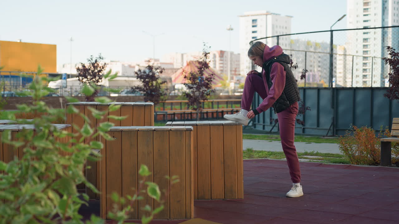 Fitness lover places foot on wooden box lifts leg high for stretching lowers it repeats with grounded stance under bright outdoor light in urban park workout session wearing hoodie vest