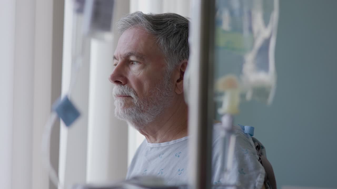 A solemn senior adult hooked up to an IV solution stands by his hospital room window framed between the plastic vacuum bags that are part of the intravenous delivery system.