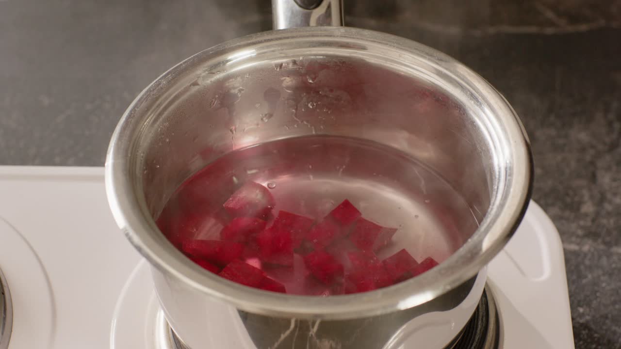 Mano afroamericana che lascia cadere cubetti rossi in una pentola che sobbolle sul fornello, mescolando il colore nell'acqua.