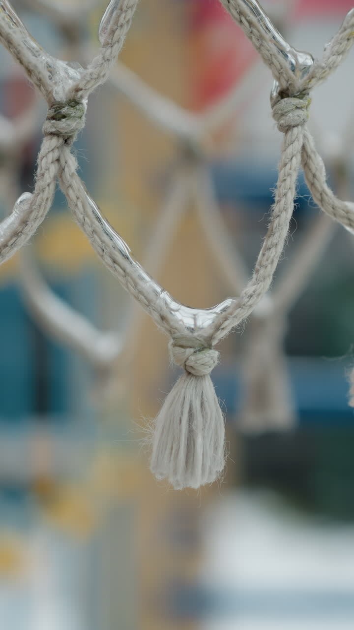 Nahaufnahme eines gefrorenen Basketballnetzes, das die eisigen Fasern und Knoten vor einem unscharfen, verschneiten Spielplatzhintergrund zeigt und den frostigen Einfluss des Winters auf Outdoor-Sportgeräte eindrucksvoll hervorhebt.