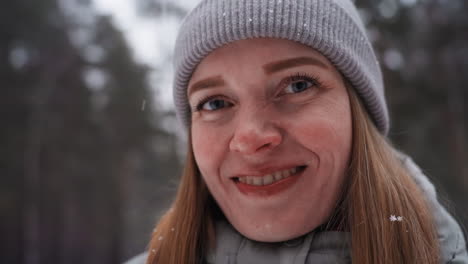 close-up portrait of smiling woman in gray beanie and winter jacket with snowflakes on hair, standing outdoors in forest during snowy cold day, conveying happiness, warmth, and peaceful seasonal atmosphere
