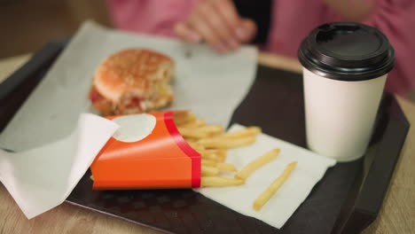 primer plano de la mano de una mujer joven en vestido rosa extiende la mano por las papas fritas de una bandeja, con una hamburguesa, papas fritas y taza de café cerca de una servilleta y comió tomó el café también y bebió de él