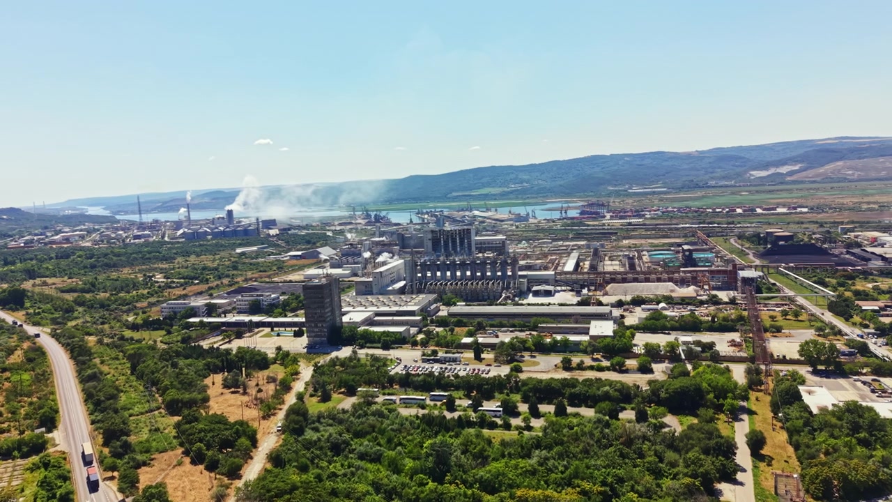 Instalación industrial ubicada cerca de un río con humo saliendo de las chimeneas y varios vehículos en la carretera durante un día luminoso