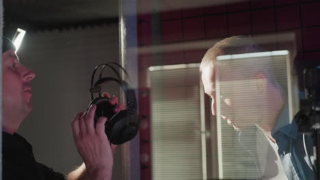 a man in a black shirt assists another in a blue long-sleeve shirt by placing headphones on him in a red soundproof studio, preparing for a recording session with a professional, collaborative