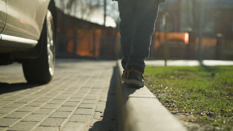 a close-up of a child carefully balancing while walking along a pavement edge next to a car, dressed in a shiny black jacket, with blurred view of trees and buildings in the background