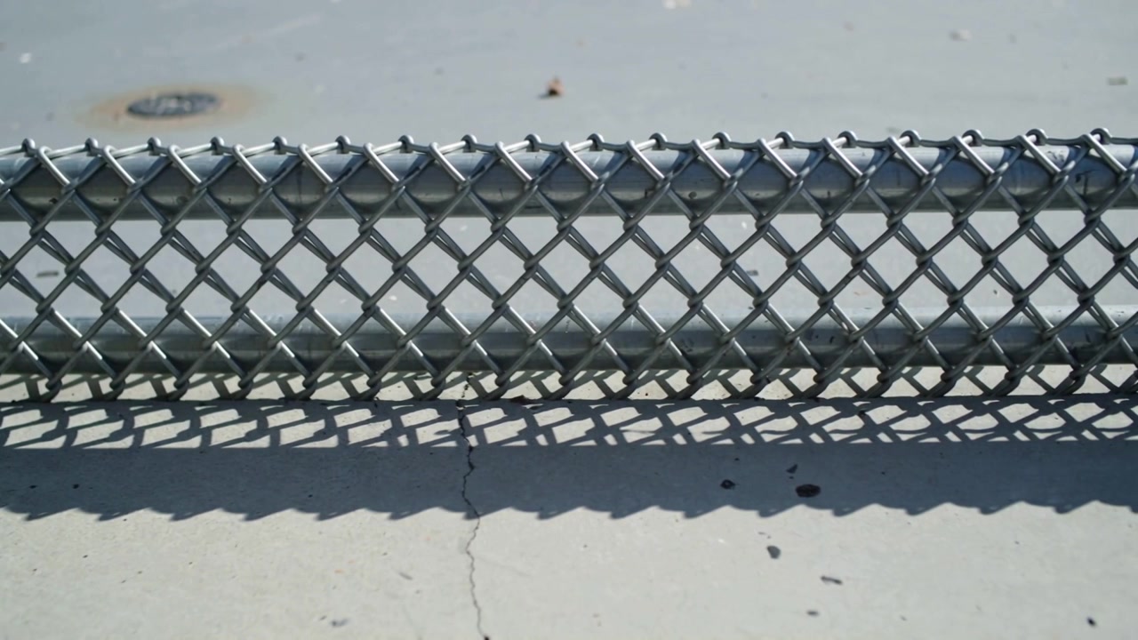 Recinzione a maglie di catena che proietta ombre a forma di diamante che si spostano su una lastra di cemento, con la luce del sole che cambia.