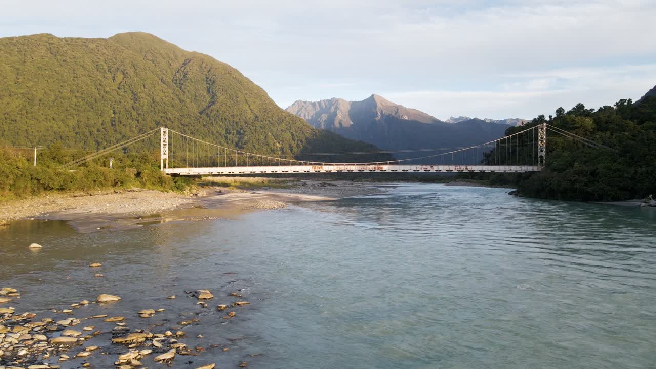 Rusty old bridge crossing blue glacial stream before rugged mountains