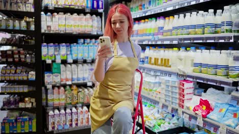 happy girl with pink hair supermarket worker takes a selfie using a white smartphone during a break from work in the dairy department in the supermarket