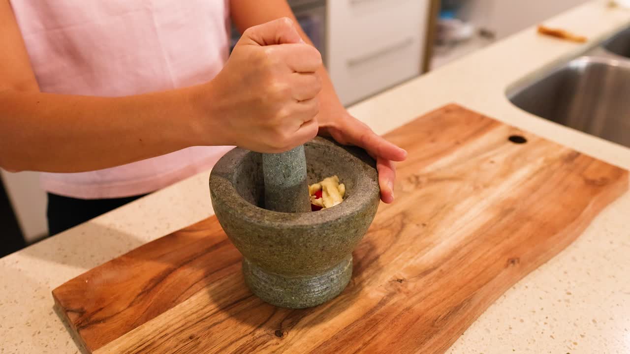 A person grinds ingredients with a mortar and pestle on a wooden board in a well-lit kitchen