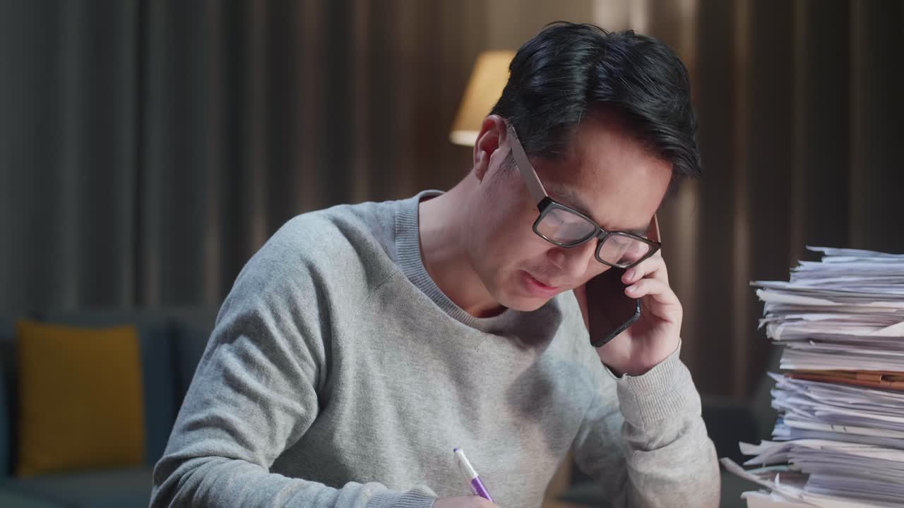 집에서 문서와 함께 열심히 일하는 동안 전화로 이야기하고 메모를 취하는 아시아 남자의 클로즈업