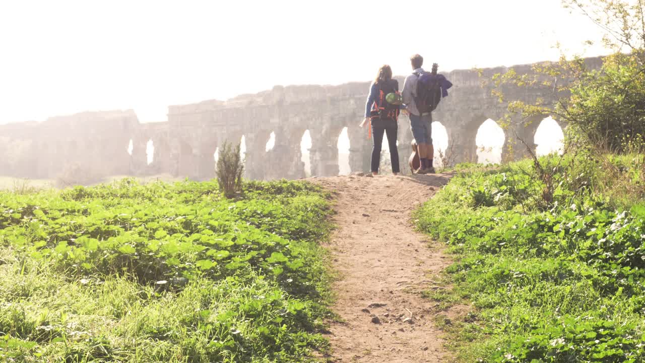 joven pareja encantadora mochileros turistas leyendo mapa señalando direcciones acueducto romano arcos en parco degli aquedotti parque ruinas en roma en romántico amanecer brumoso con guitarra y saco de dormir cámara lenta