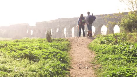 joven pareja encantadora mochileros turistas leyendo mapa señalando direcciones acueducto romano arcos en parco degli aquedotti parque ruinas en roma en romántico amanecer brumoso con guitarra y saco de dormir cámara lenta
