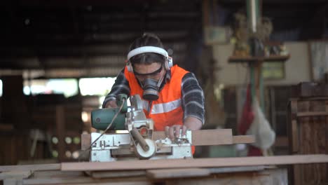 carpenter using circular saw cutting wooden plank in workshop . young man builder sawing board . craftsman wearing protective mask , hearing protectors and safety glasses .