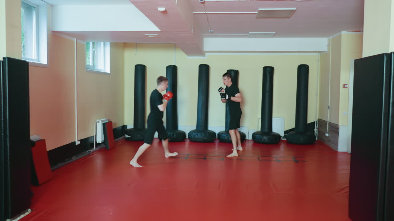 무술 체육관 안에서 선수들이 스파링하는 모습이에요. 한 선수는 빨간 글러브를 끼고 공격적인 자세로 앞으로 나아가고, 상대 선수는 빨간 매트 바닥에서 검은색 펀칭 백을 배경으로 방어 태세를 취하고 있어요.