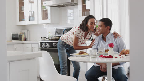 Hombre-Afroamericano-Milenario-Sentado-En-La-Cocina-Mientras-Su-Pareja-Les-Sirve-Una-Comida-Romántica