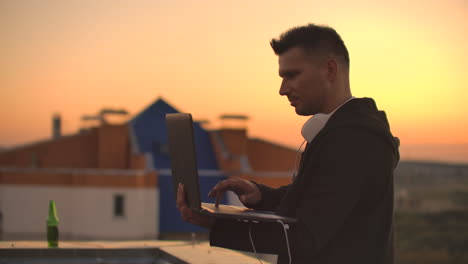 a young man sitting on the edge of the roof with a laptop and a beer working typing on a laptop